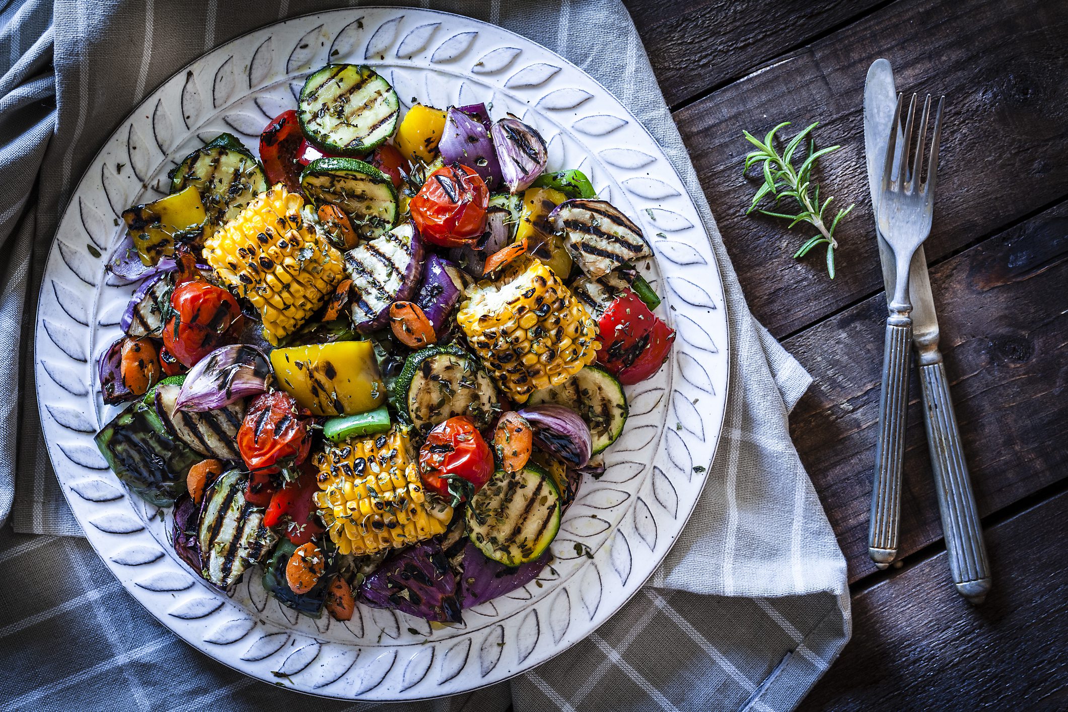 Close-up view of a grilled vegetables plate shot from above on rustic wooden kitchen table. The plate is on a gray textile napkin at the left of an horizontal frame and a fork and knife are at the right of the plate. DSRL studio photo taken with Canon EOS 5D Mk II and Canon EF 100mm f/2.8L Macro IS USM